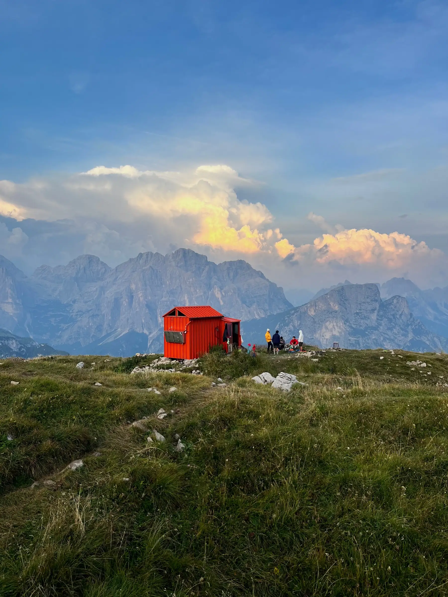 A red bivouac hut on a grass ridge, Dolomite walls and a sunlit cloud behind it