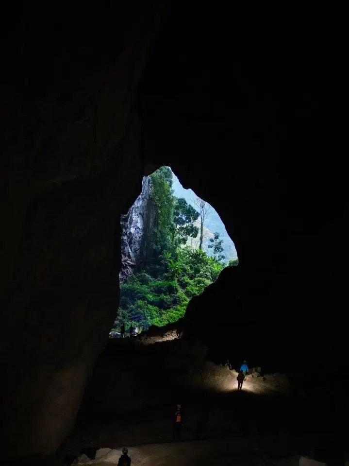 Small figures with headtorches inside a vast cave mouth, jungle light beyond it
