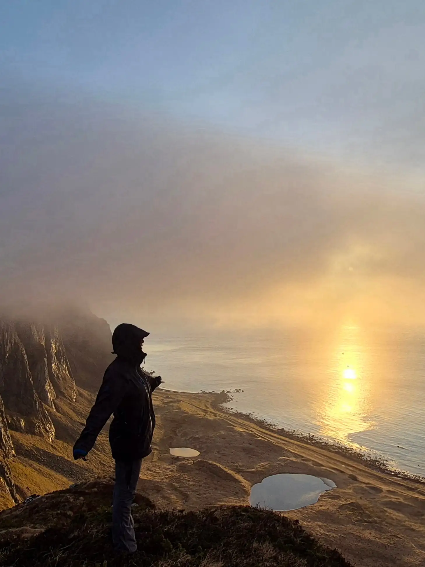 A figure on a cliff edge above the sea, a low sun burning through the mist
