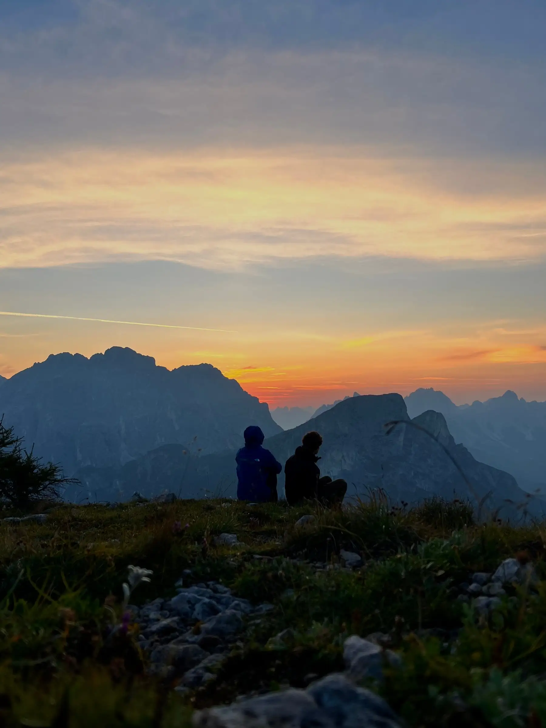 Two people sitting on a grass ridge, watching the sun go down behind the Dolomites