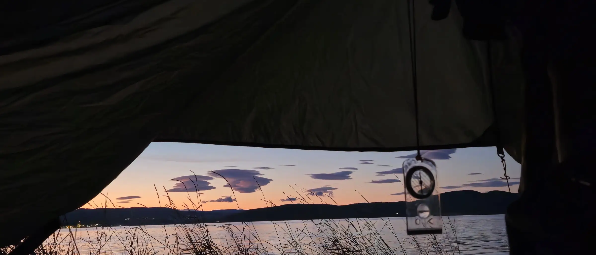 Looking out of a tent at dusk across a lake, lenticular clouds above the far shore and a compass hanging in the doorway